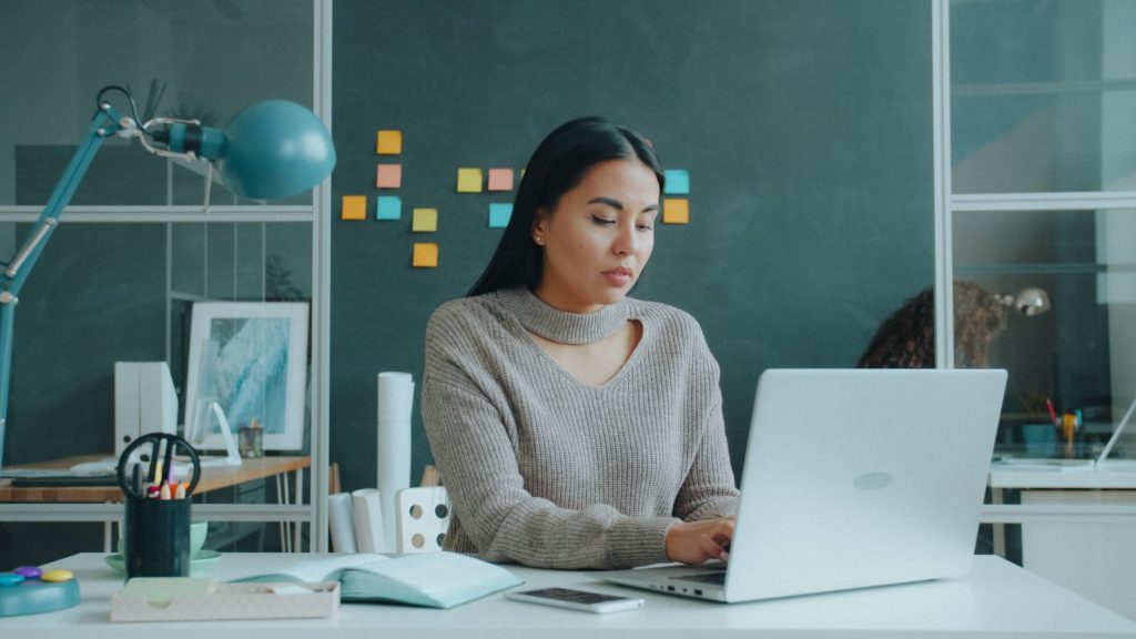 A woman using a laptop in a modern office setting with notebooks and sticky notes on the wall.